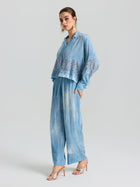 A woman wears Social Queen's Washed Silk Wide-Leg Pants with an elastic drawstring waist, paired with a loose, light blue lace blouse, heeled sandals, and large earrings, standing against a plain background.