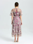 A woman faces away, wearing the Social Queen Floral Beaded A-Line Dress with sheer flutter sleeves. Her brown hair is in a low updo, and she pairs the look with heels.