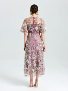 A woman faces away, wearing the Social Queen Floral Beaded A-Line Dress with sheer flutter sleeves. Her brown hair is in a low updo, and she pairs the look with heels.