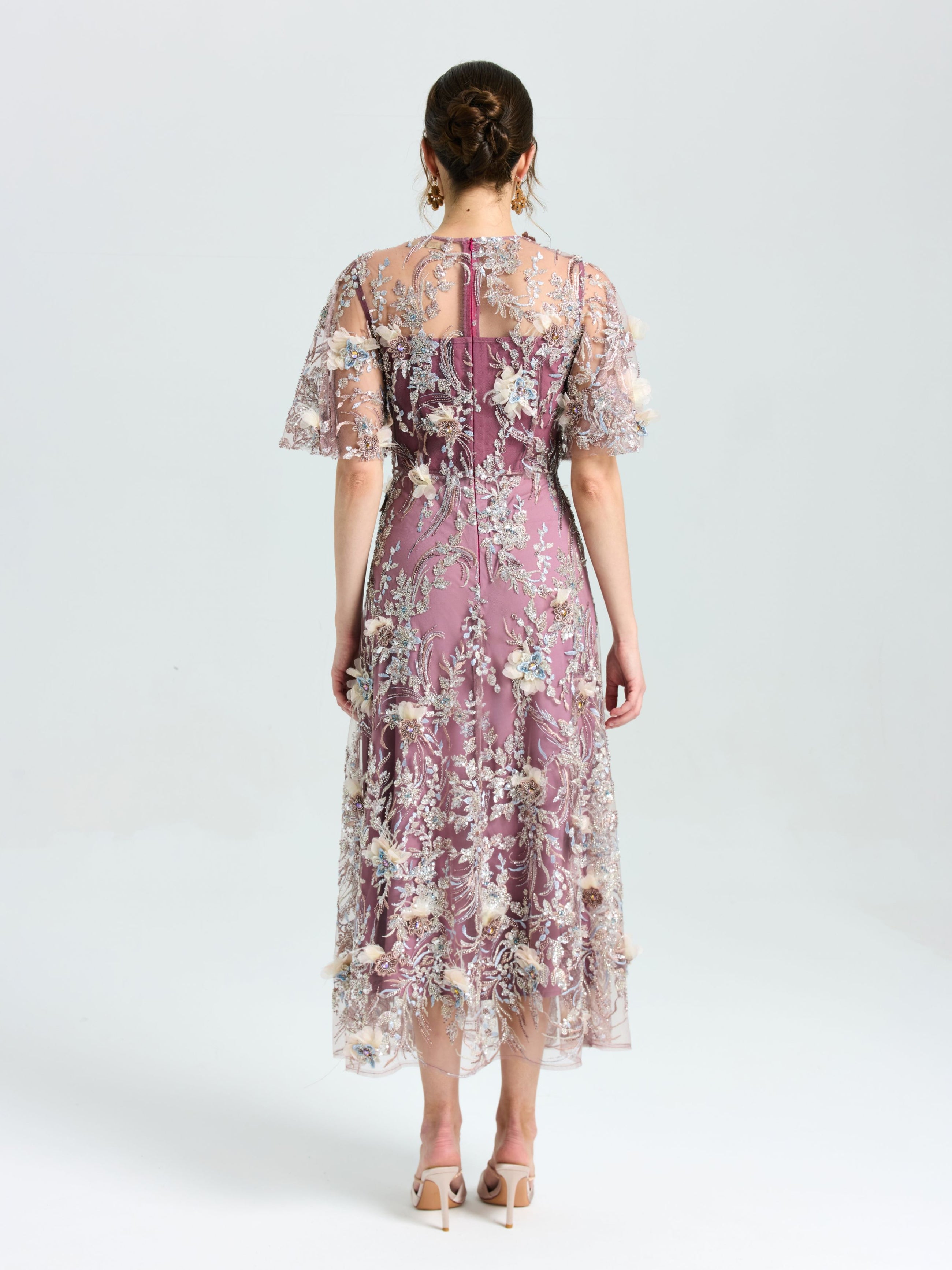 A woman faces away, wearing the Social Queen Floral Beaded A-Line Dress with sheer flutter sleeves. Her brown hair is in a low updo, and she pairs the look with heels.