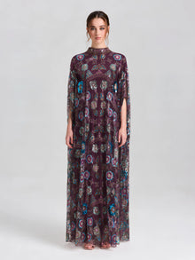 A woman poses in a studio wearing the Social Queen Beaded Floral Embellished Cape Gown—an elegant long, sheer purple gown with floral sequins and short sleeves—set against a neutral background.