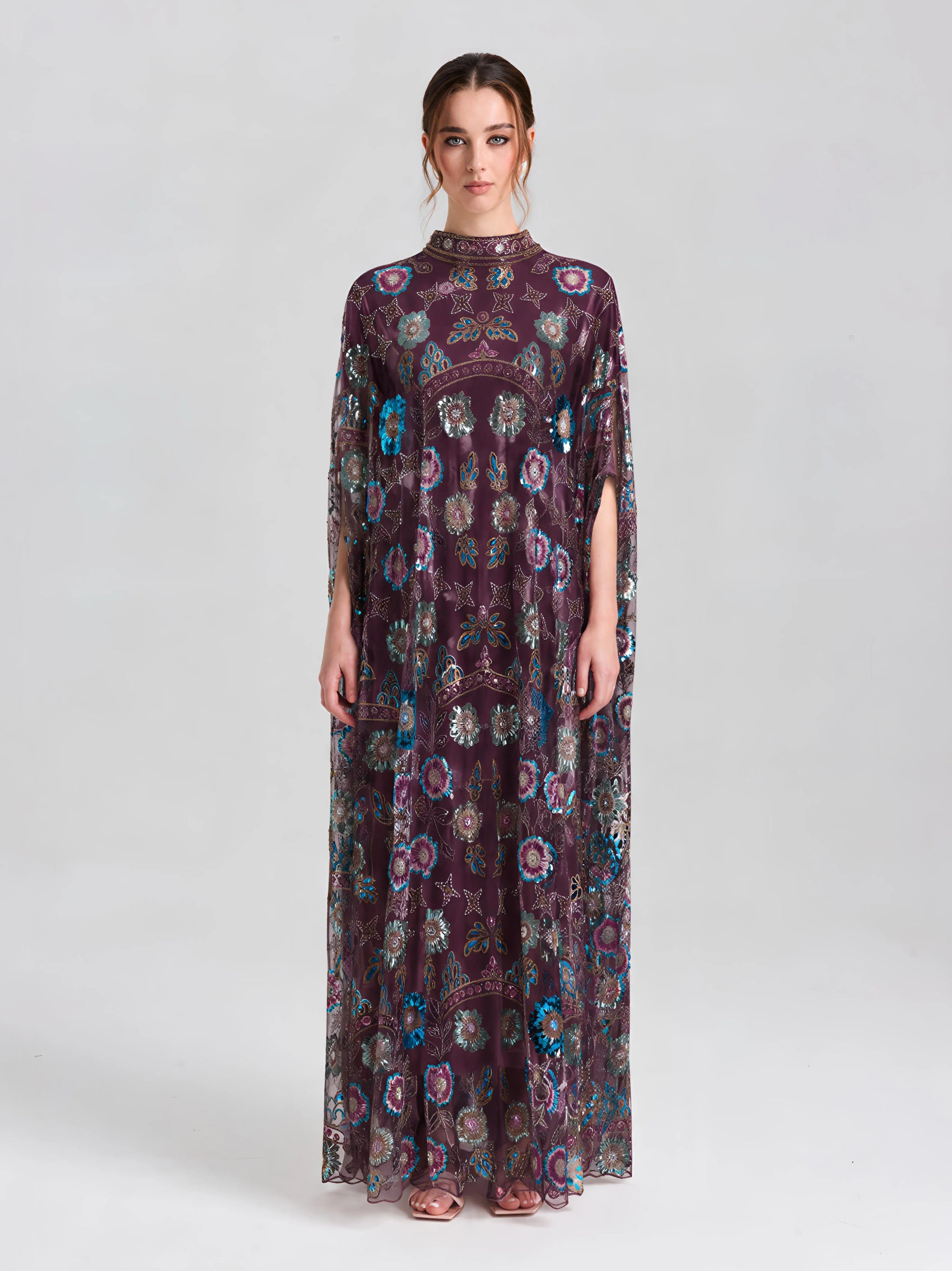 A woman poses in a studio wearing the Social Queen Beaded Floral Embellished Cape Gown—an elegant long, sheer purple gown with floral sequins and short sleeves—set against a neutral background.