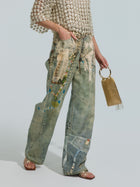 Wearing a textured white top, the person sports Social Queen’s Pearl Chain Printed Jeans with a wide-leg silhouette and artistic painted designs, paired with metallic sandals and carrying a small gold mesh handbag.