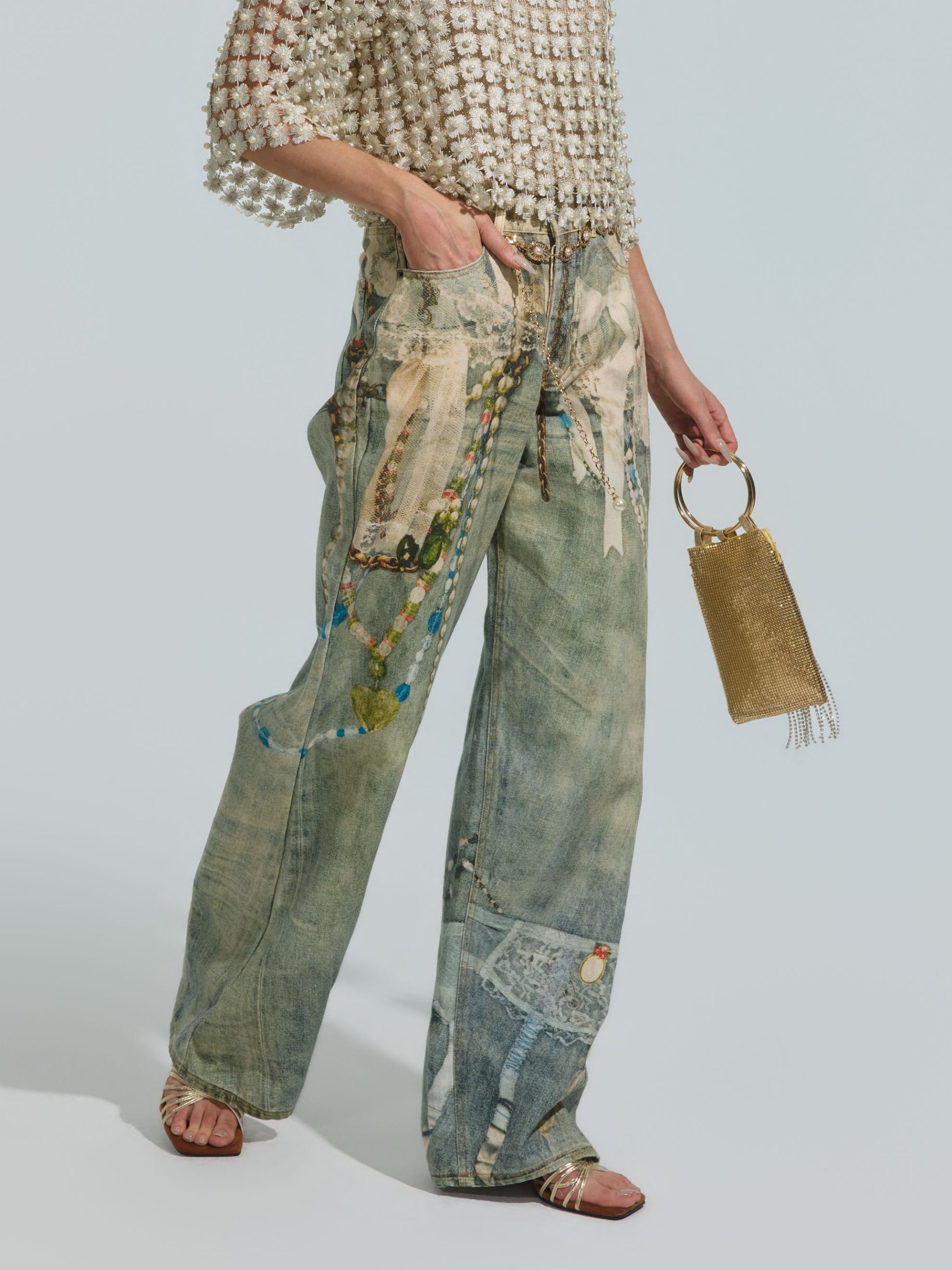 Wearing a textured white top, the person sports Social Queen’s Pearl Chain Printed Jeans with a wide-leg silhouette and artistic painted designs, paired with metallic sandals and carrying a small gold mesh handbag.