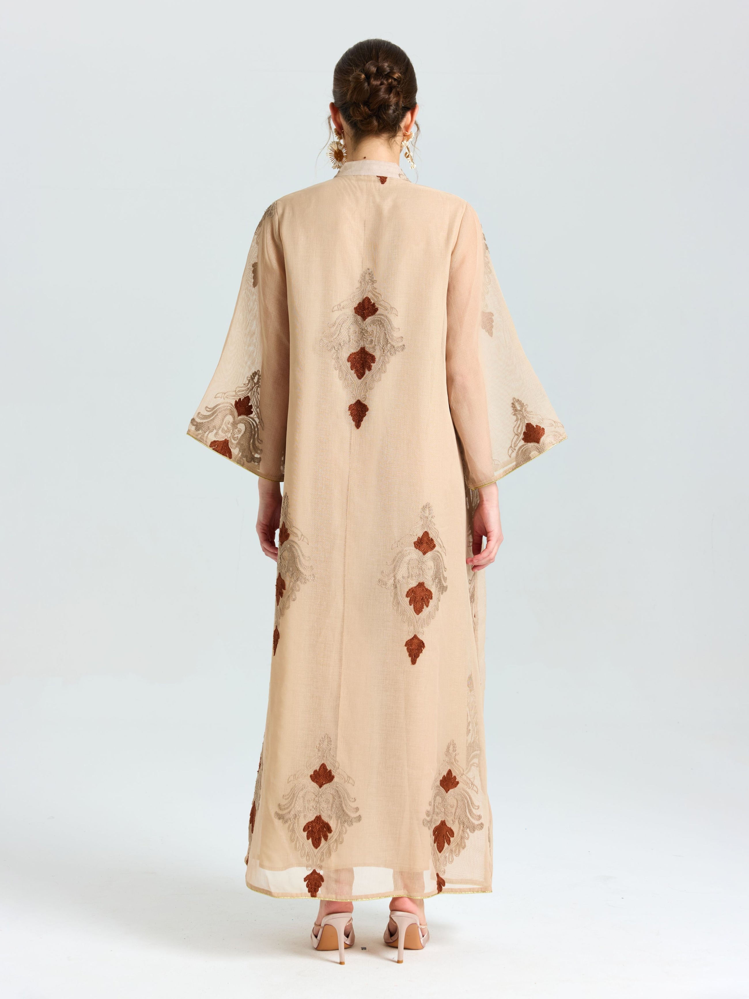 A person stands facing away, wearing the Social Queen Placement Embroidered Linen Dress with wide sleeves and brown damask scroll embroidery, paired with high-heeled sandals.