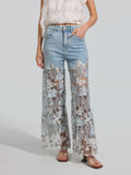 A person wearing Social Queen’s Floral Lace Patchwork Pants—light blue stretch denim wide-leg pants with sheer floral lace patchwork panels from mid-thigh to hem—paired with a white floral top and sandals, stands against a plain background.