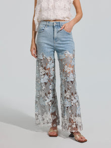 A person wearing Social Queen’s Floral Lace Patchwork Pants—light blue stretch denim wide-leg pants with sheer floral lace patchwork panels from mid-thigh to hem—paired with a white floral top and sandals, stands against a plain background.