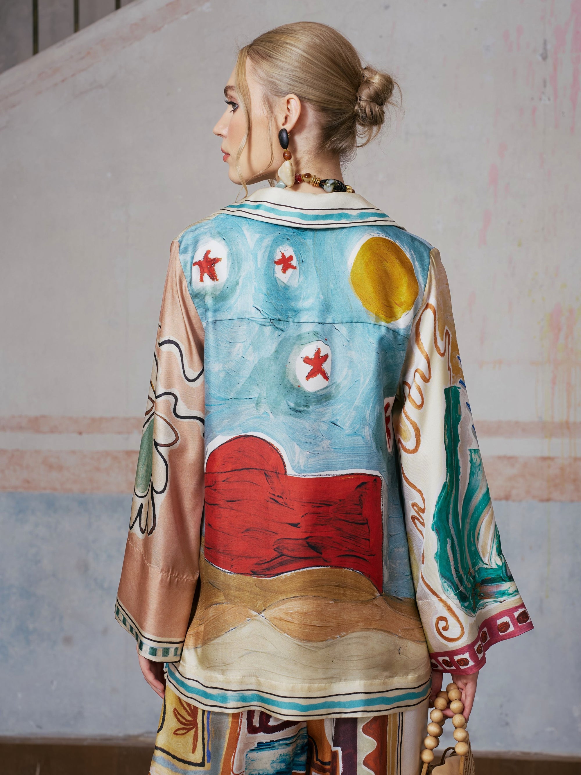 A person with blonde hair in a low bun wears the Social Queen Abstract Print Mulberry Silk Shirt, standing with their back to the camera against a light, textured wall.