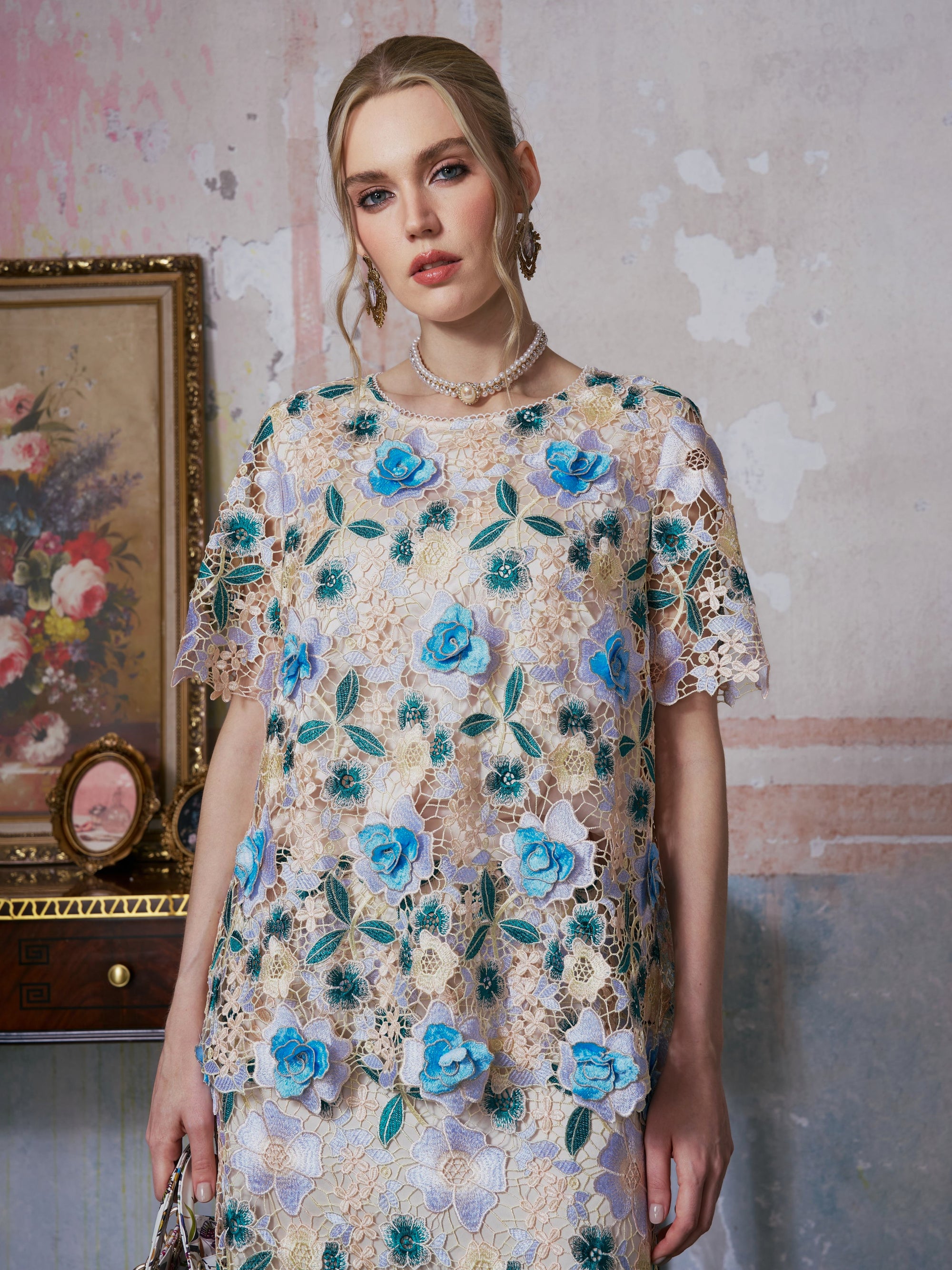 Wearing the 3D Floral Cutwork Lace Top by Social Queen, a woman stands indoors near a framed floral painting and a vintage table.