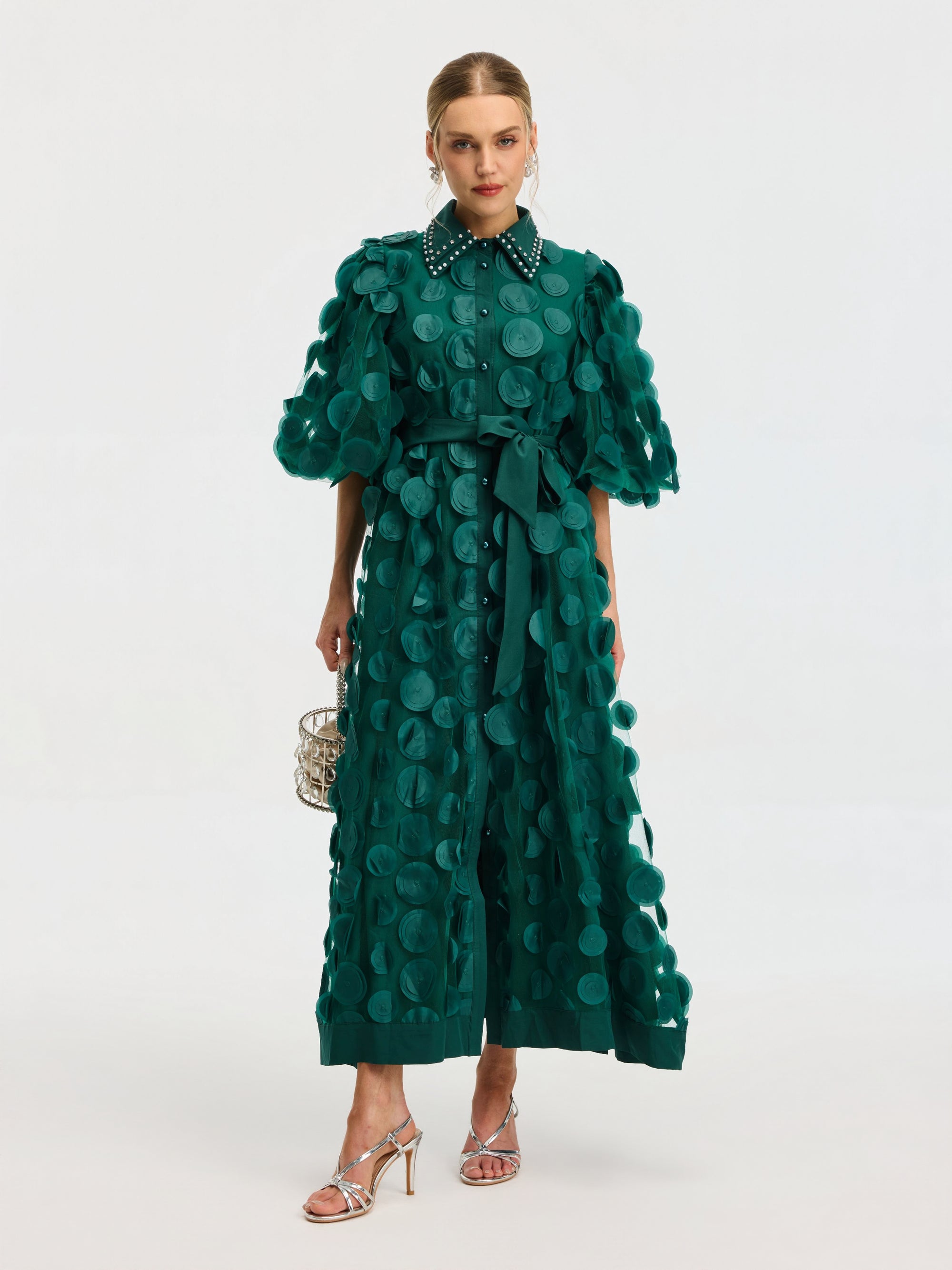 A woman wears the Social Queen Pearl Embellished Appliqué Dress, featuring a green textured polka dot pattern, belted waist, and pearl collar. She carries a metallic handbag and pairs the look with silver heels.