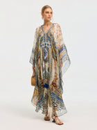 A woman in the Social Queen Byzantine Print Silk Caftan Dress stands against a plain light background, accessorized with a woven handbag and sandals.