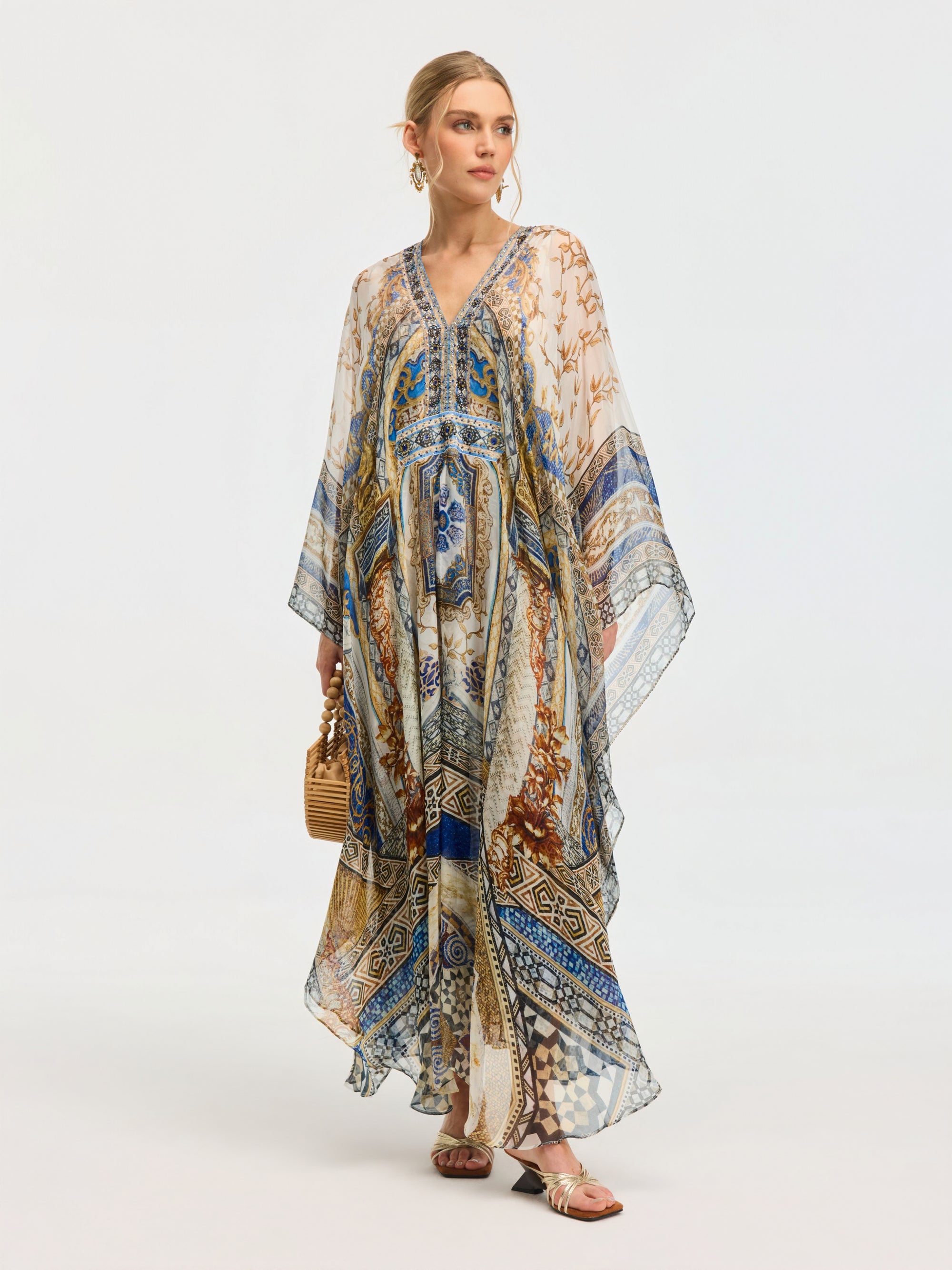 A woman in the Social Queen Byzantine Print Silk Caftan Dress stands against a plain light background, accessorized with a woven handbag and sandals.