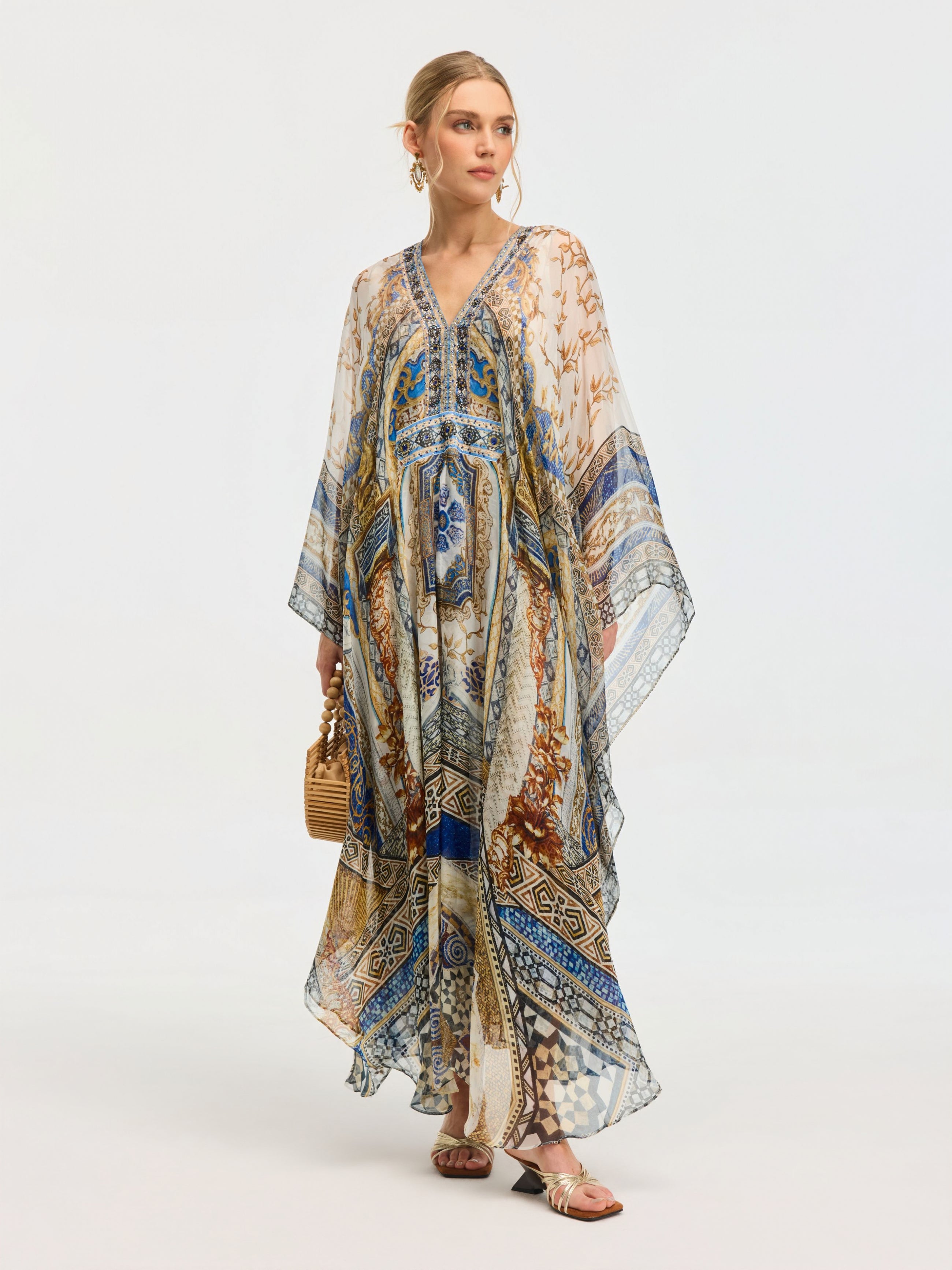 A woman in the Social Queen Byzantine Print Silk Caftan Dress stands against a plain light background, accessorized with a woven handbag and sandals.