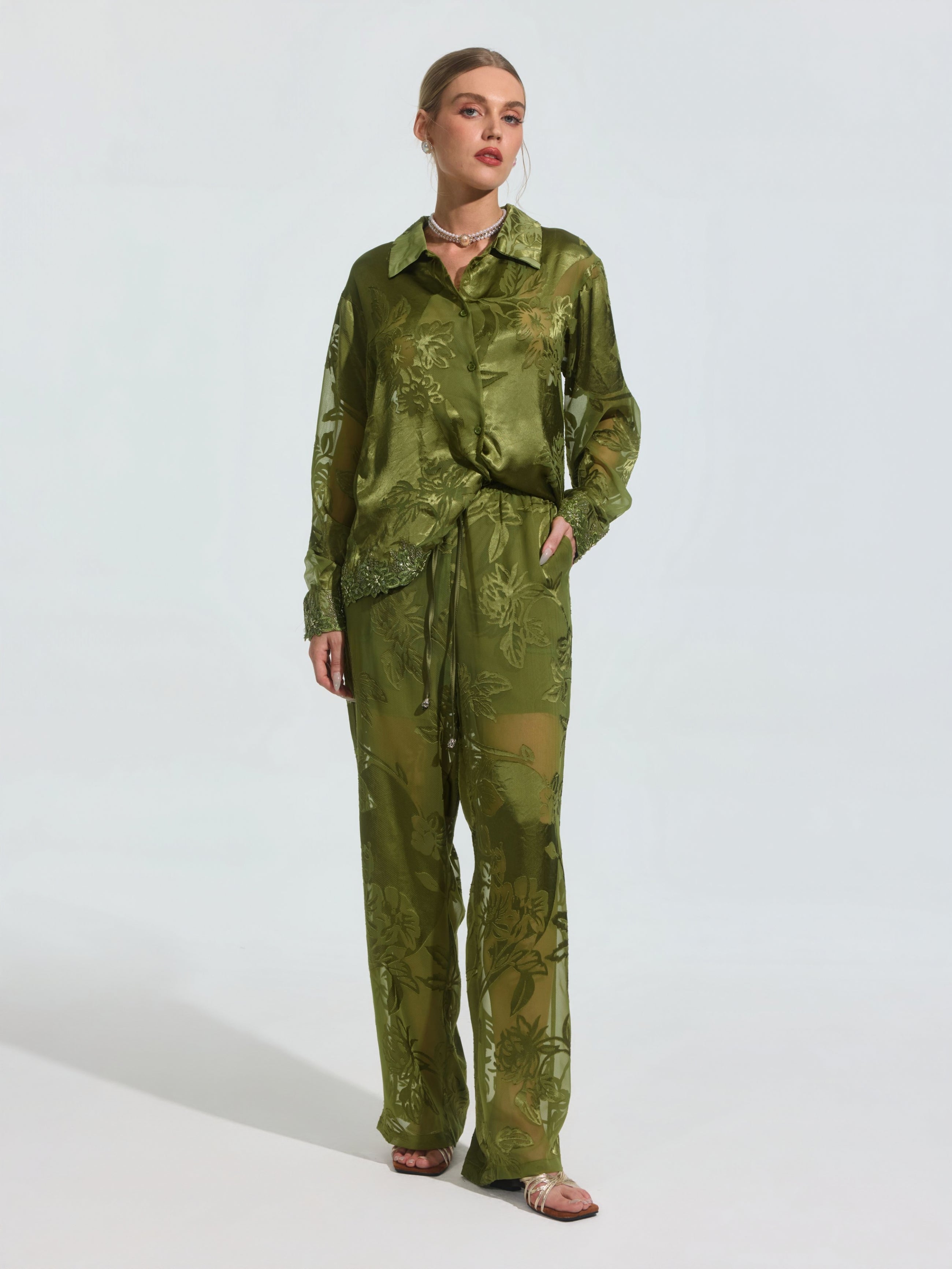 A woman wears the Social Queen Beaded Botanical Burnout Satin Shirt in olive green, featuring a floral pattern, paired with matching wide-leg pants and sandals against a plain light background.