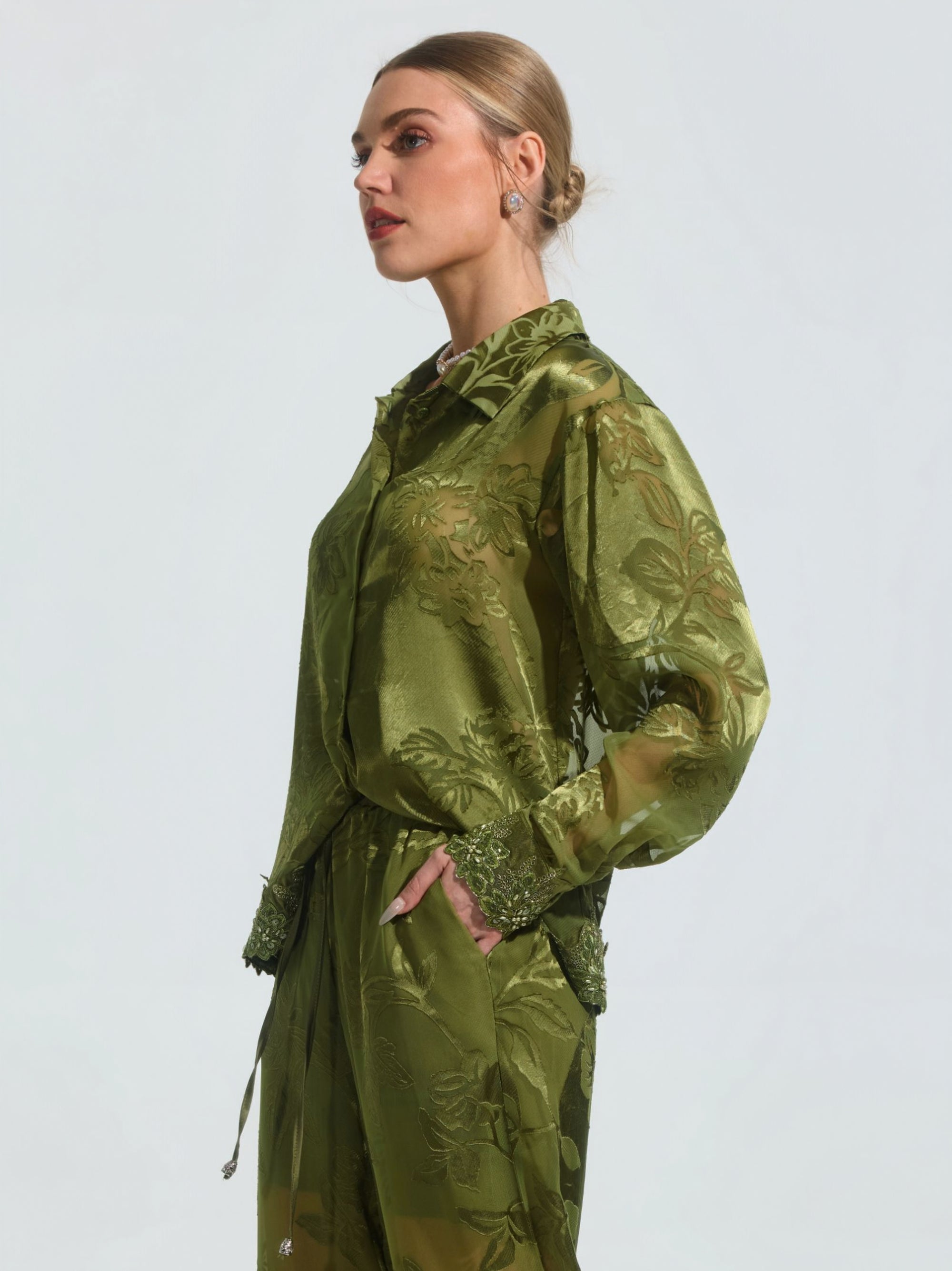 A woman stands in profile with her hand in her pocket, wearing the Social Queen Beaded Botanical Burnout Satin Shirt—a sheer green top featuring a floral pattern and beaded lace trim—against a plain light background.