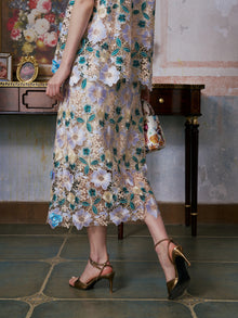 Wearing the Social Queen 3D Floral Cutwork Lace Skirt and gold heels, a person stands indoors holding a small decorative handbag, with part of a vintage desk in the background.