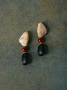 Social Queen Multicolor Beaded Drop Earrings, featuring irregular cream stones, round brown beads, and matte black oval drops, are displayed on a textured green surface.