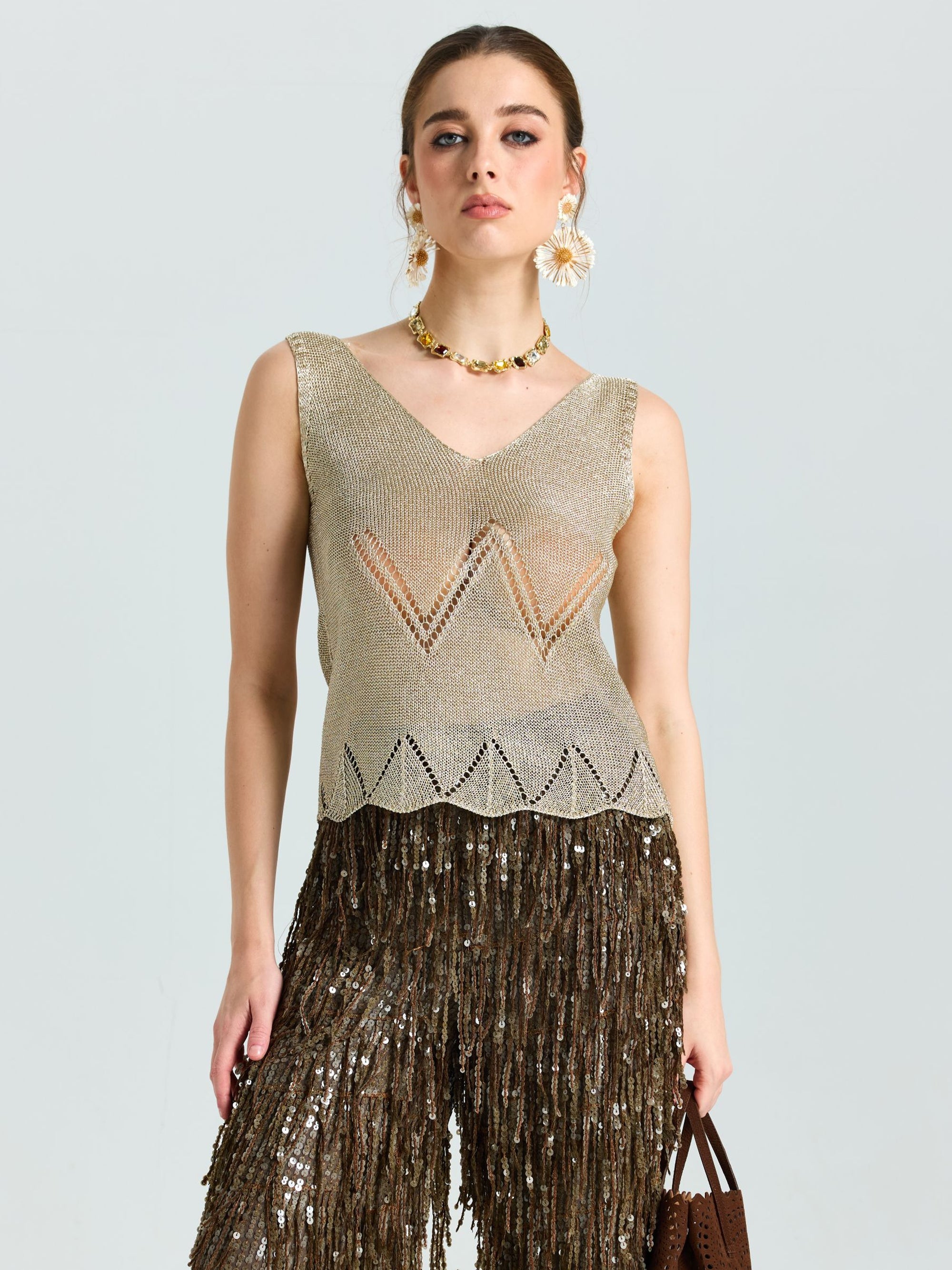 A woman in the Social Queen Open-Knit Metallic Sleeveless Top stands against a plain background, styled with geometric pants, large floral earrings, and a statement necklace.