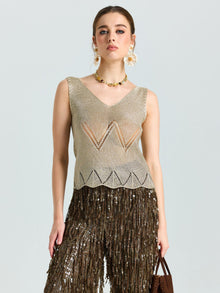 A woman in the Social Queen Open-Knit Metallic Sleeveless Top stands against a plain background, styled with geometric pants, large floral earrings, and a statement necklace.