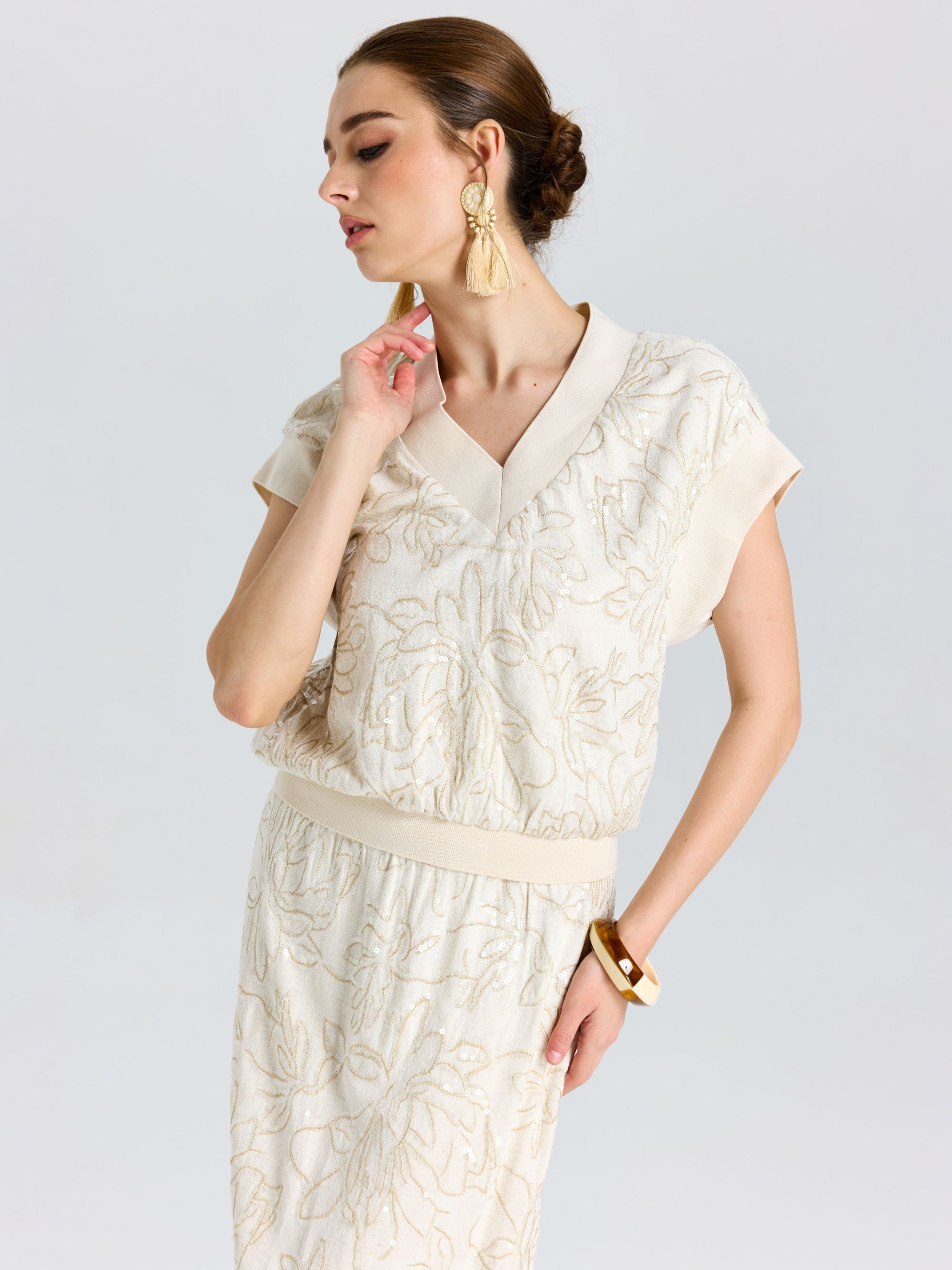 Woman in a Social Queen Floral Embroidered Linen Top with short sleeves, paired with a skirt, tassel earrings, and a bangle, standing against a plain background.
