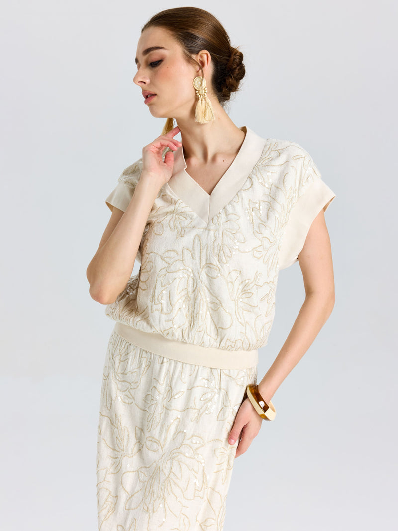 Woman in a Social Queen Floral Embroidered Linen Top with short sleeves, paired with a skirt, tassel earrings, and a bangle, standing against a plain background.