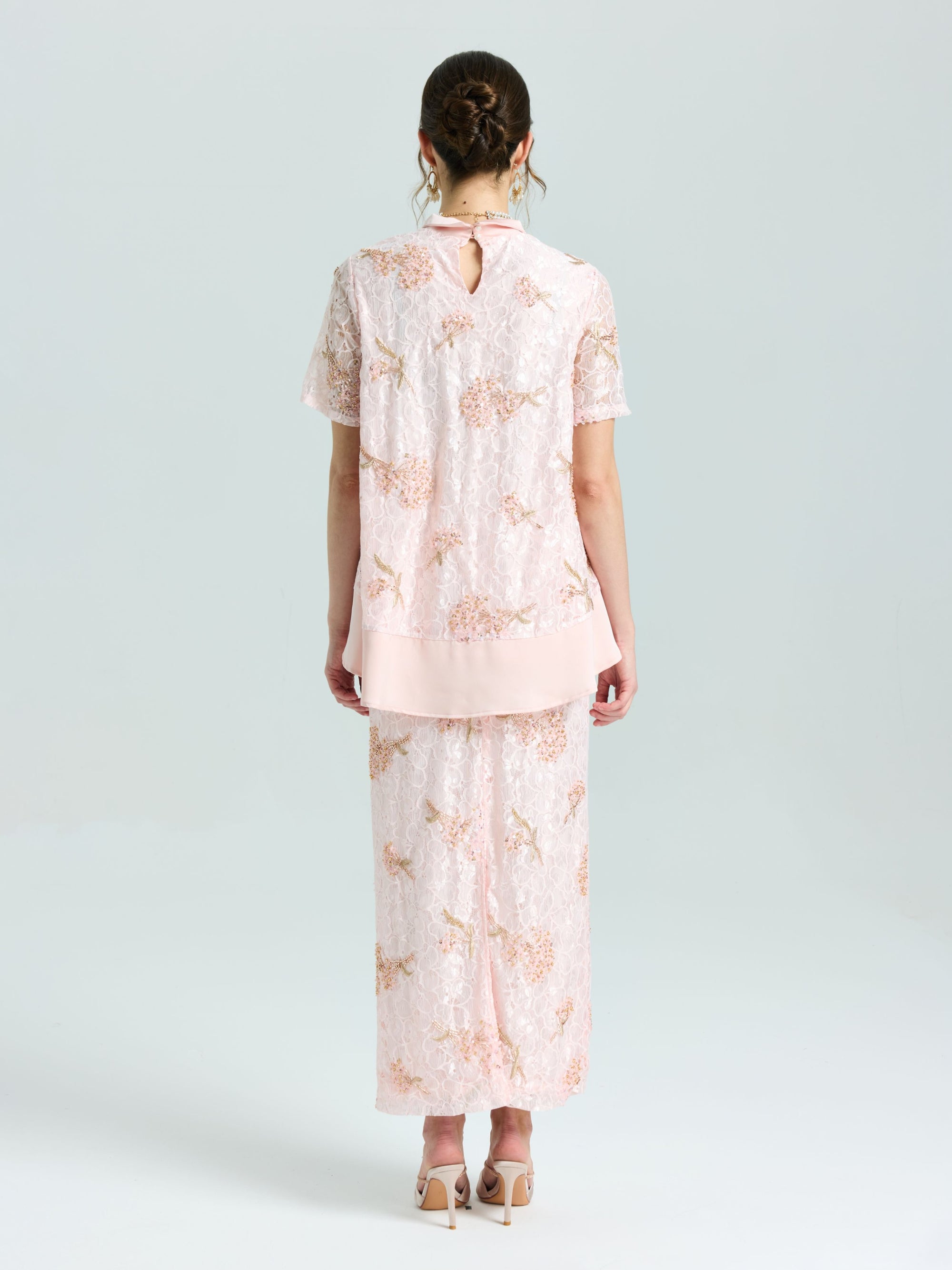 A woman stands with her back to the camera wearing Social Queen’s Dragonfly Beaded Lace Midi Skirt and matching top in light pink, both embellished with delicate beadwork, set against a simple pale background.