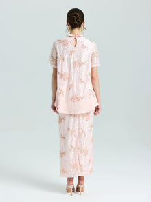 A woman stands with her back to the camera wearing Social Queen’s Dragonfly Beaded Lace Midi Skirt and matching top in light pink, both embellished with delicate beadwork, set against a simple pale background.
