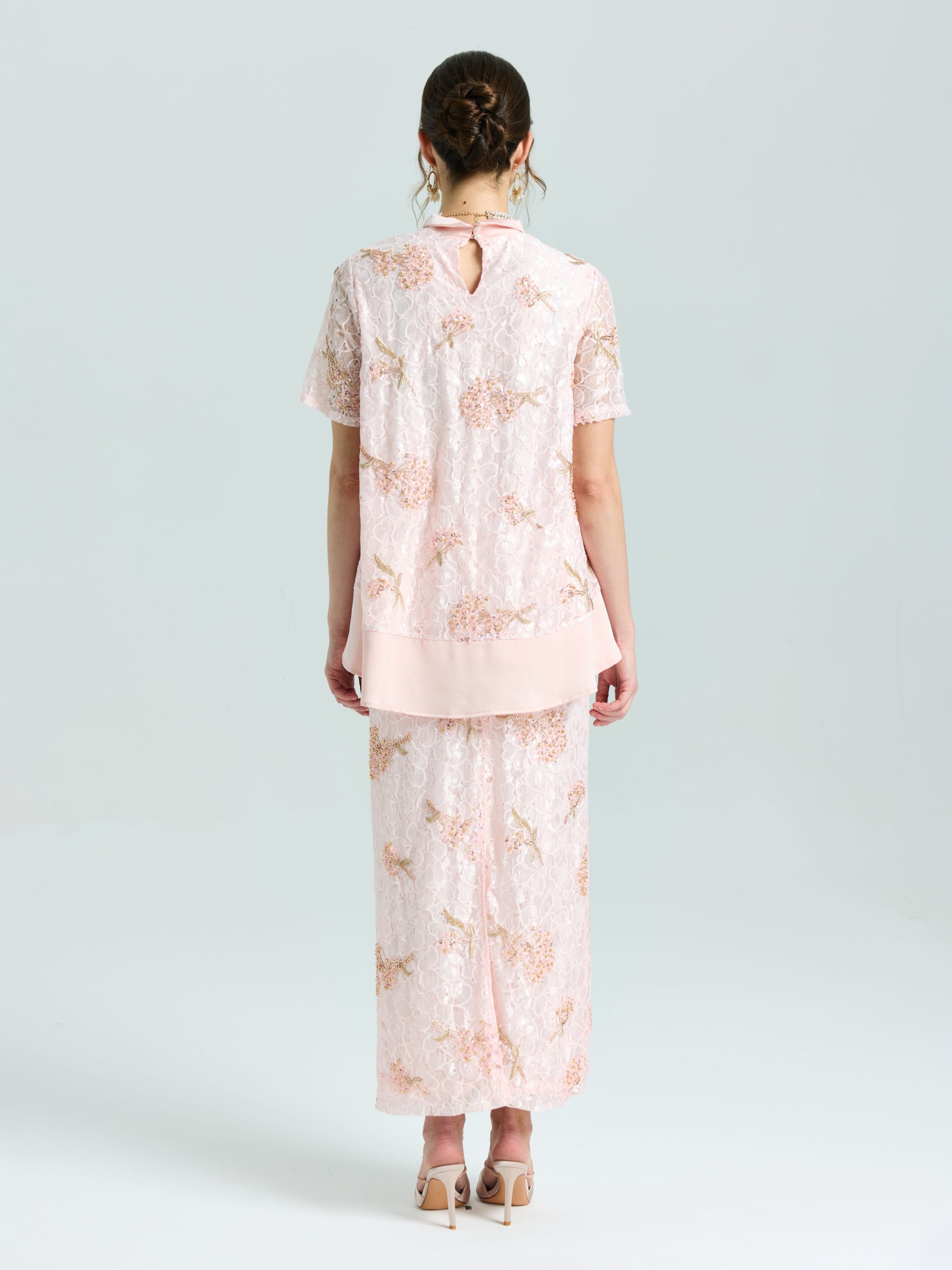 A woman stands with her back to the camera wearing Social Queen’s Dragonfly Beaded Lace Midi Skirt and matching top in light pink, both embellished with delicate beadwork, set against a simple pale background.