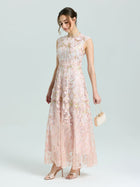 A woman in the Social Queen Gold Floral Embroidered Dress—long, sleeveless, and blush pink with delicate gold floral details—stands against a plain light background, holding a beige purse and wearing open-toe heels.