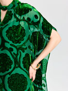 A person wearing the Social Queen Floral Velvet Cape Dress (Cami Included), featuring sheer green silk velvet with dark green floral burnout patterns, a subtle cape-inspired silhouette, and accessorized with a gold bracelet.