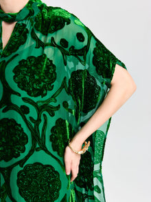A person wearing the Social Queen Floral Velvet Cape Dress (Cami Included), featuring sheer green silk velvet with dark green floral burnout patterns, a subtle cape-inspired silhouette, and accessorized with a gold bracelet.