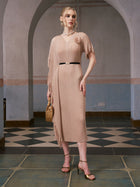 A woman stands indoors in front of an arched wall and tiled floor, wearing the Social Queen Asymmetric Fringe Midi Dress with included black belt, gold heels, and holding a woven purse.