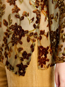 Close-up of a person wearing the Social Queen Flocked Floral Silk Sheer Top (Camisole Not Included), featuring metallic vine-inspired embroidery, paired with tan velvet pants. Only the torso and arm are visible.