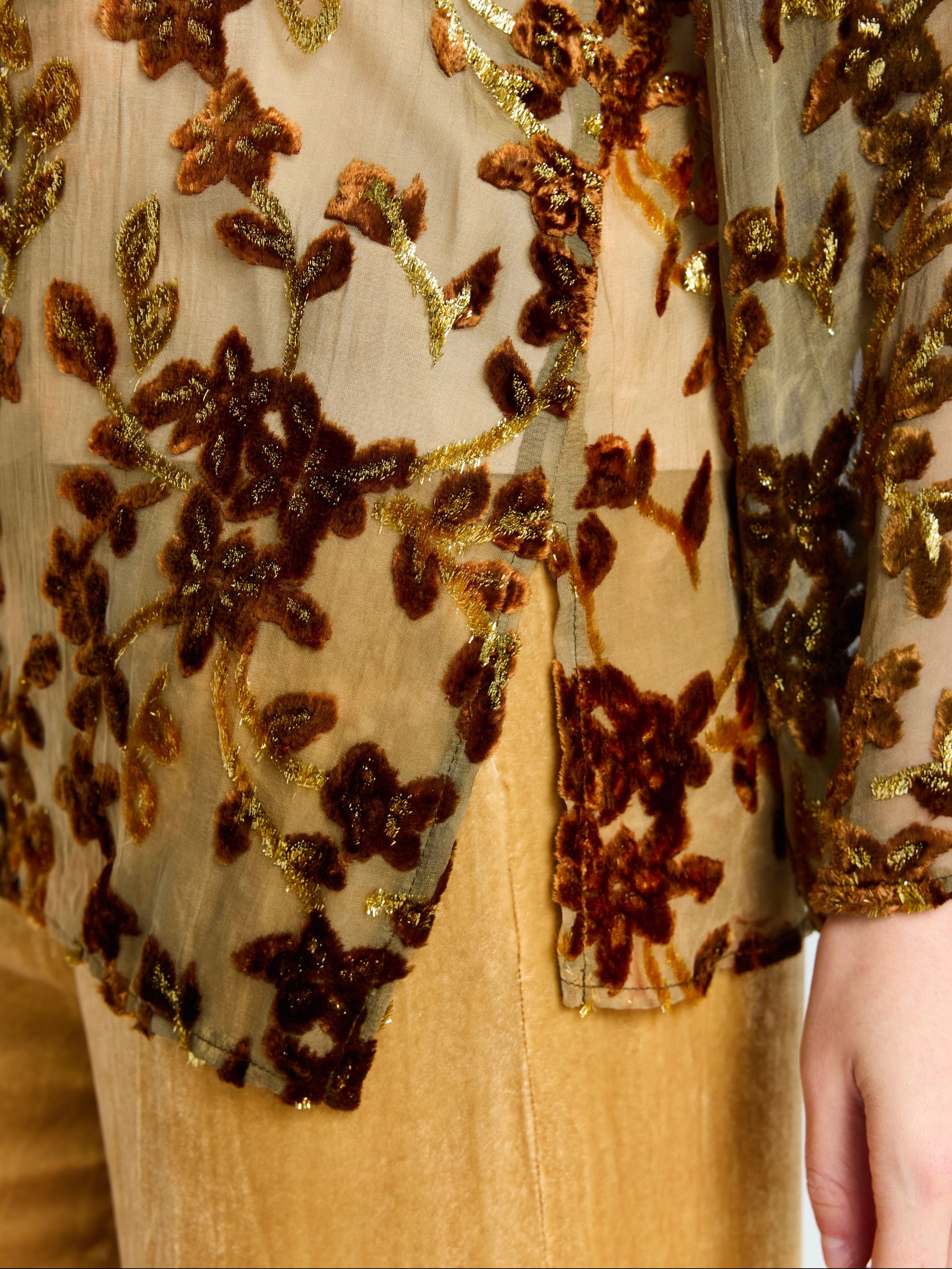 Close-up of a person wearing the Social Queen Flocked Floral Silk Sheer Top (Camisole Not Included), featuring metallic vine-inspired embroidery, paired with tan velvet pants. Only the torso and arm are visible.