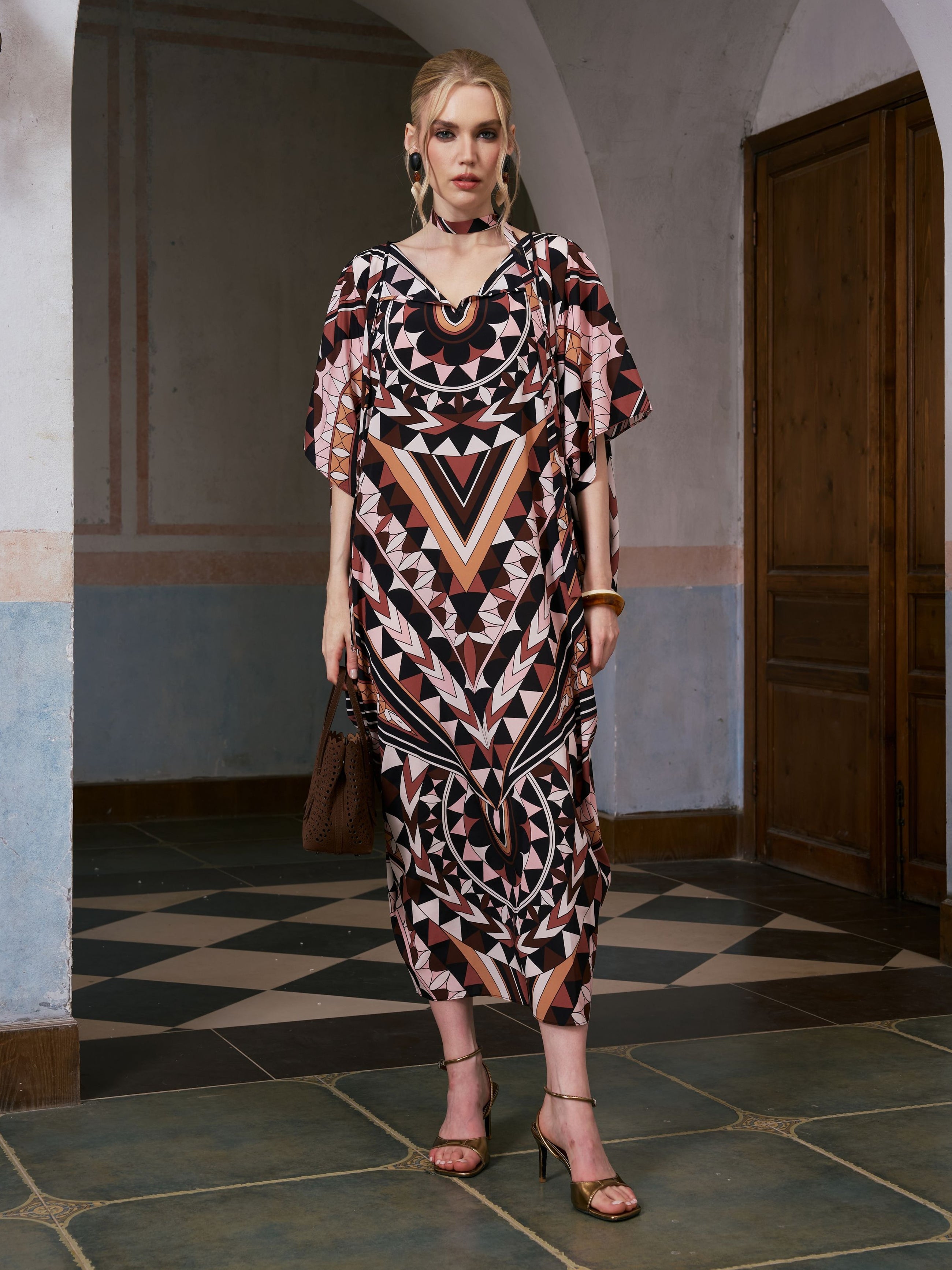 A woman stands indoors wearing the Social Queen Geometric Motif Print Silk Dress (Scarf Included) paired with heeled sandals and holding a brown bag. Tiled flooring, an arched doorway, and muted wall colors in the background evoke a sense of relaxed resort wear elegance.