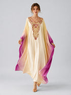 A woman wears the Social Queen Ombré Beaded Embroidered Cape Dress—a long satin gown with cream-to-purple ombré coloring and intricate chest embroidery—paired with gold sandals, standing against a plain light gray background.