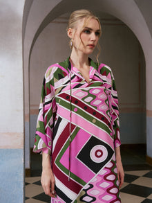 A woman wearing the Social Queen Geometric Print Mulberry Silk Dress, featuring a robe-style silhouette and vibrant geometric patterns, stands in an arched hallway and gazes to the side with a neutral expression.