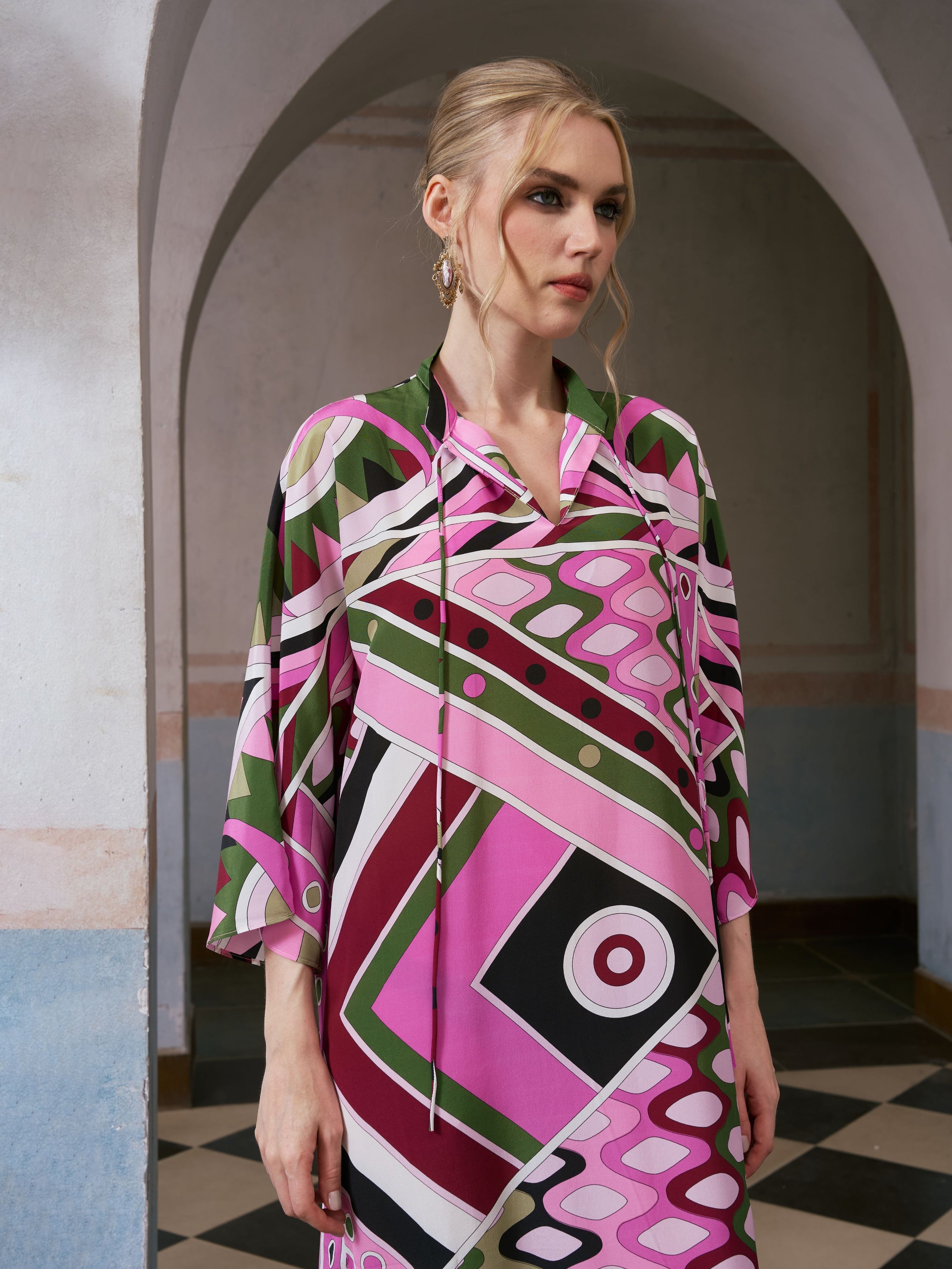A woman wearing the Social Queen Geometric Print Mulberry Silk Dress, featuring a robe-style silhouette and vibrant geometric patterns, stands in an arched hallway and gazes to the side with a neutral expression.