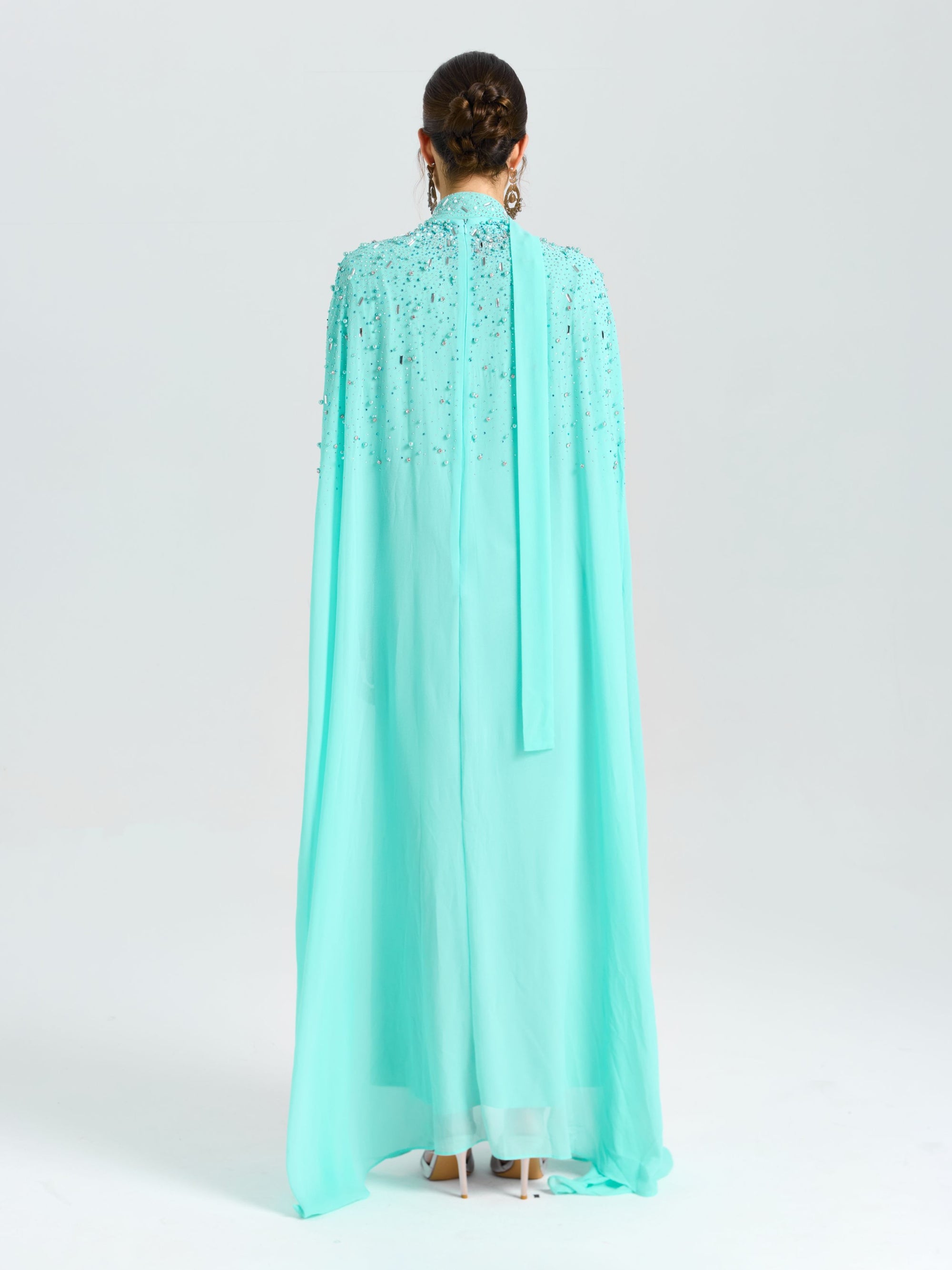 A person faces away from the camera in a Social Queen Beaded Cape Scarf Gown with earrings, set against a plain light background.