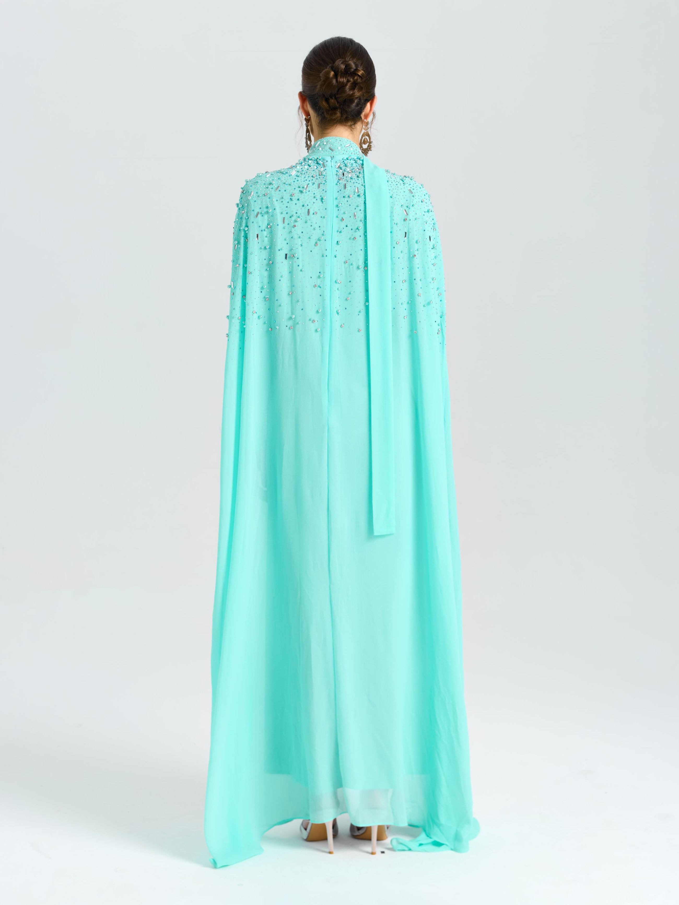 A person faces away from the camera in a Social Queen Beaded Cape Scarf Gown with earrings, set against a plain light background.