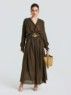 A woman models the Social Queen Twist-Front Ramie Shirt, featuring long sleeves and a twist-front waist, paired with a tan handbag and open-toe heels against a plain light background.