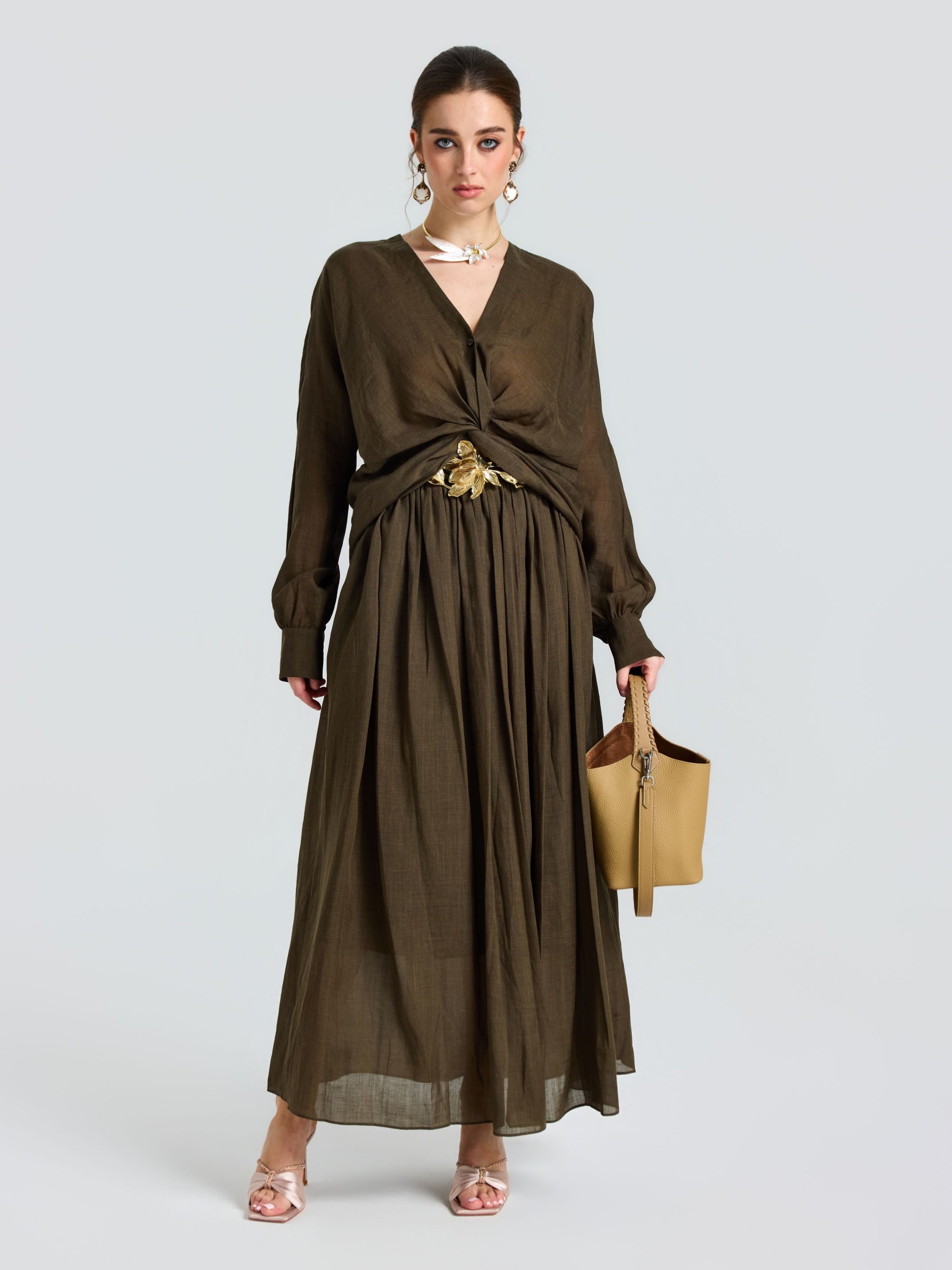 A woman models the Social Queen Twist-Front Ramie Shirt, featuring long sleeves and a twist-front waist, paired with a tan handbag and open-toe heels against a plain light background.