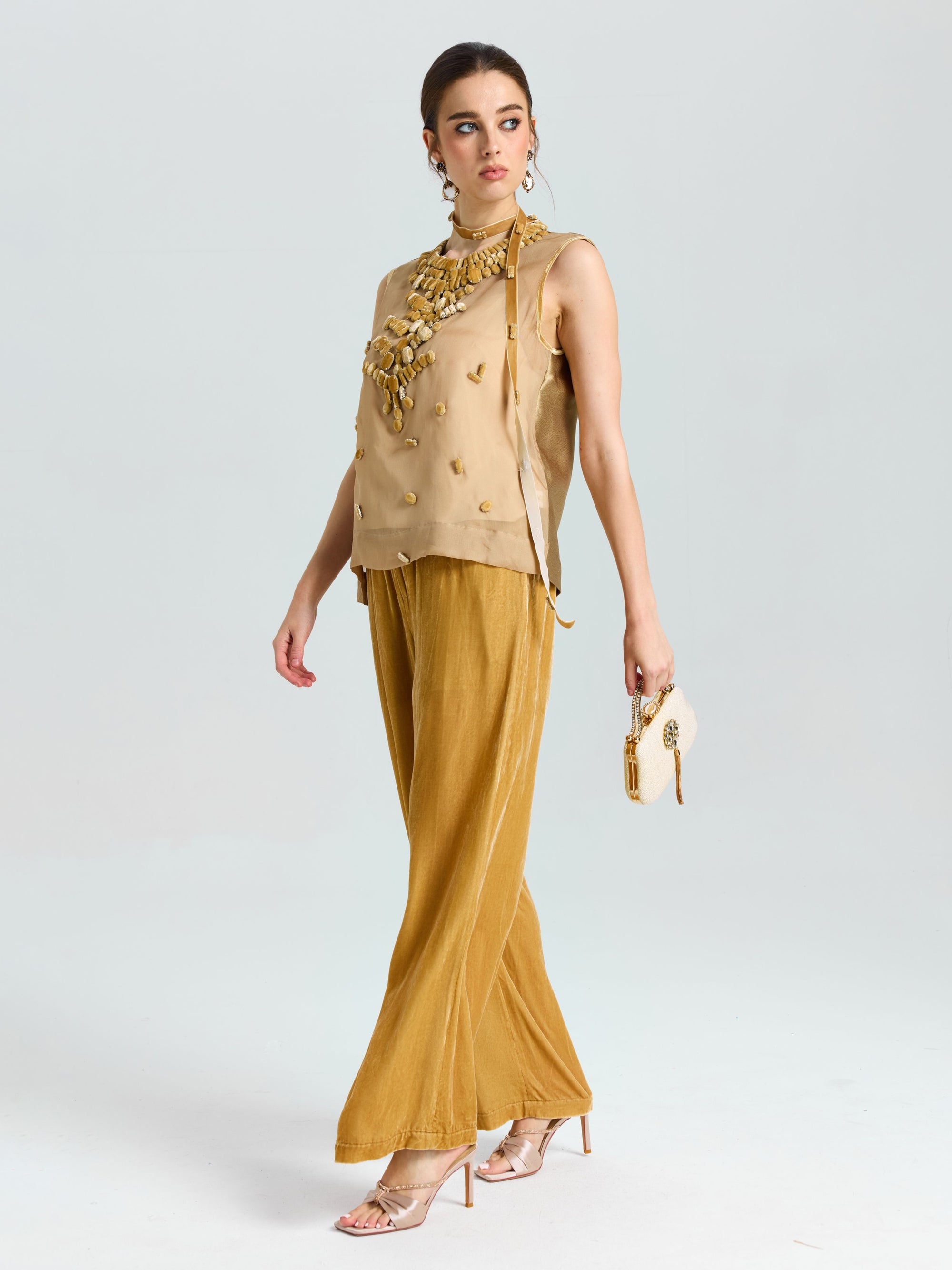 A woman in the Social Queen Velvet Scarf Silk Organza Top (cami included), wide-legged gold pants, and heels holds a small clutch against a plain light background.