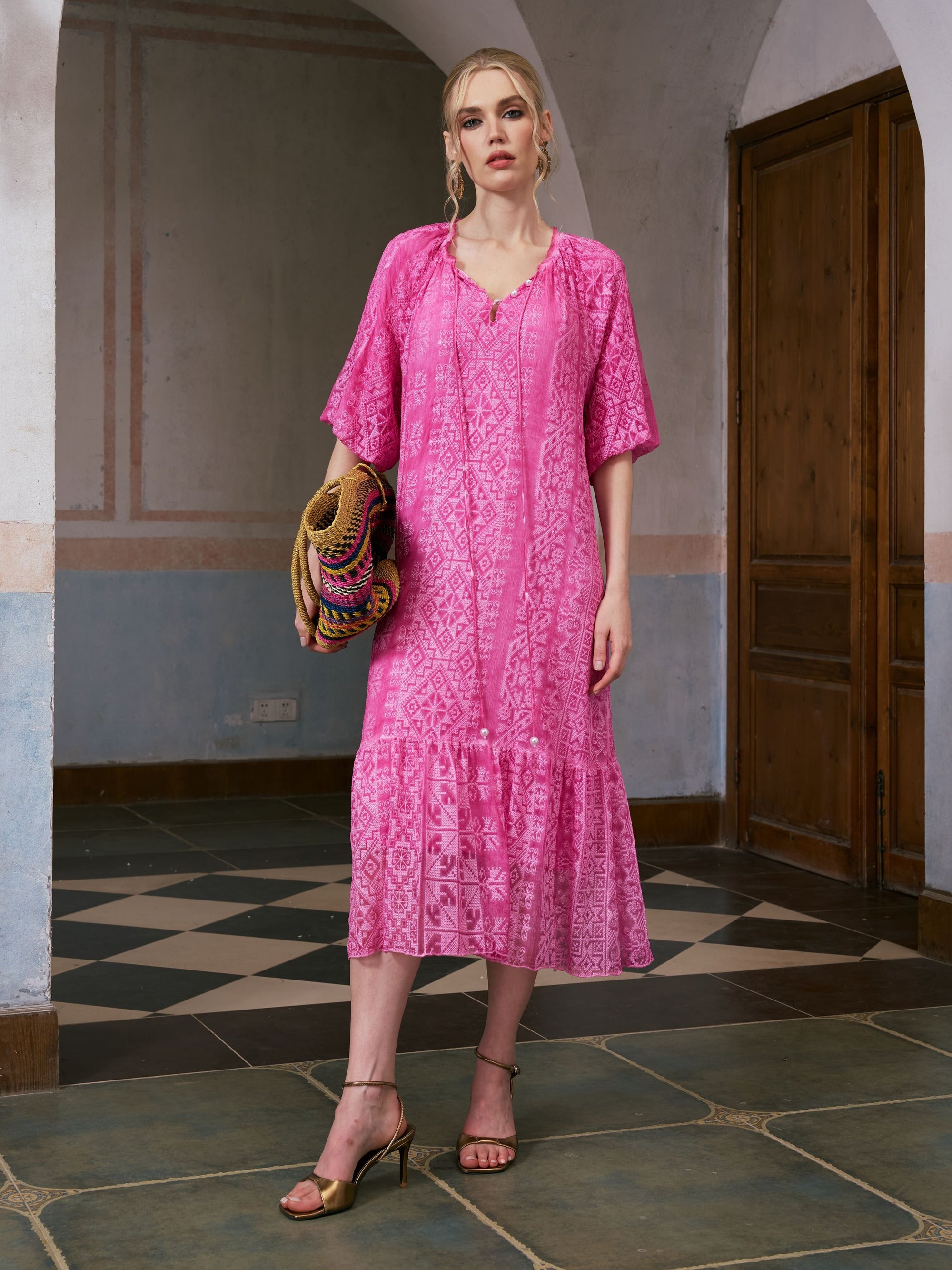 A woman stands indoors near a wooden door, wearing the Social Queen Geometric Jacquard Mulberry Silk Dress with tan sandals and carrying a colorful woven bag on a tiled floor—an ideal look for effortless resort wear.