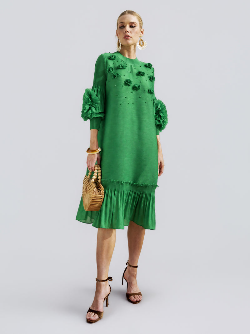 A woman stands against a plain background wearing the Social Queen Beaded Floral Pleated Midi Dress, featuring ruffled sleeves and hem. She accessorizes with a wooden handbag, gold earrings, and black high-heeled sandals.