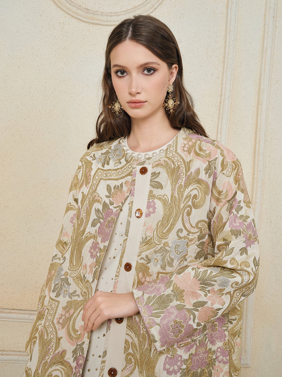 A woman with long brown hair wears the Social Queen Italian Baroque Floral Metallic Jacquard Jacket with large buttons and ornate earrings, standing against a textured cream-colored wall.