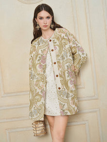 A woman stands against a beige wall wearing the Social Queen Italian Baroque Floral Metallic Jacquard Jacket over a white dress with gold studs, holding a beaded handbag.