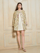 A woman stands indoors wearing the Social Queen Italian Baroque Floral Metallic Jacquard Jacket over a white dress with gold accents, paired with a small gold handbag and gold sandals.