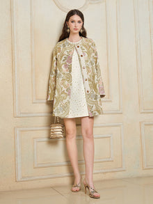 A woman stands indoors wearing the Social Queen Italian Baroque Floral Metallic Jacquard Jacket over a white dress with gold accents, paired with a small gold handbag and gold sandals.