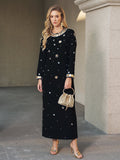 A woman stands on a stone floor in the Social Queen Jewel-Beaded Gold-Trim Dress with gold sandals, a woven handbag, and jewelry.